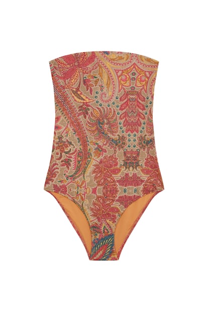Modelo desfilando con bañador one‑piece de corte bandeau‑sirena y rayas verticales en lurex, sobre fondo de playa soleada