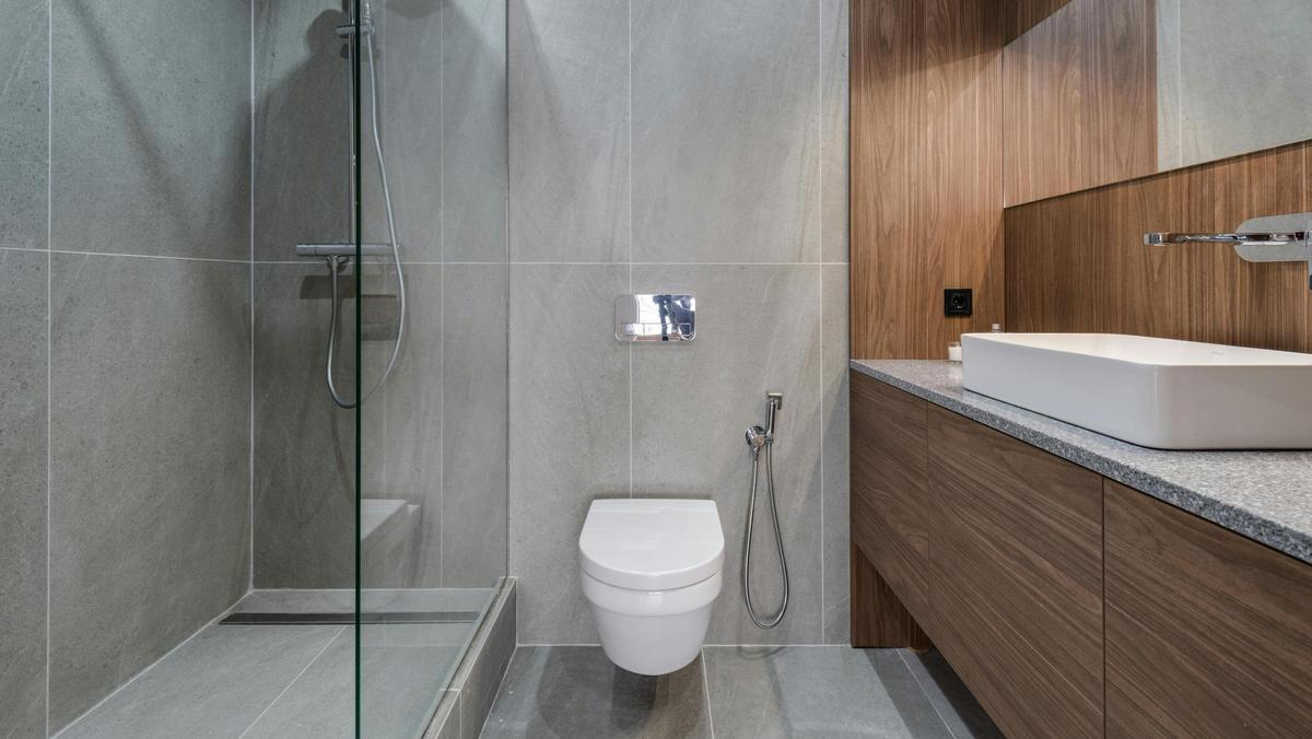 Baño reformado con lavadora y secadora integradas en la ducha, mostrando cerámica lisa y en relieve, inodoro suspendido y hornacina de madera